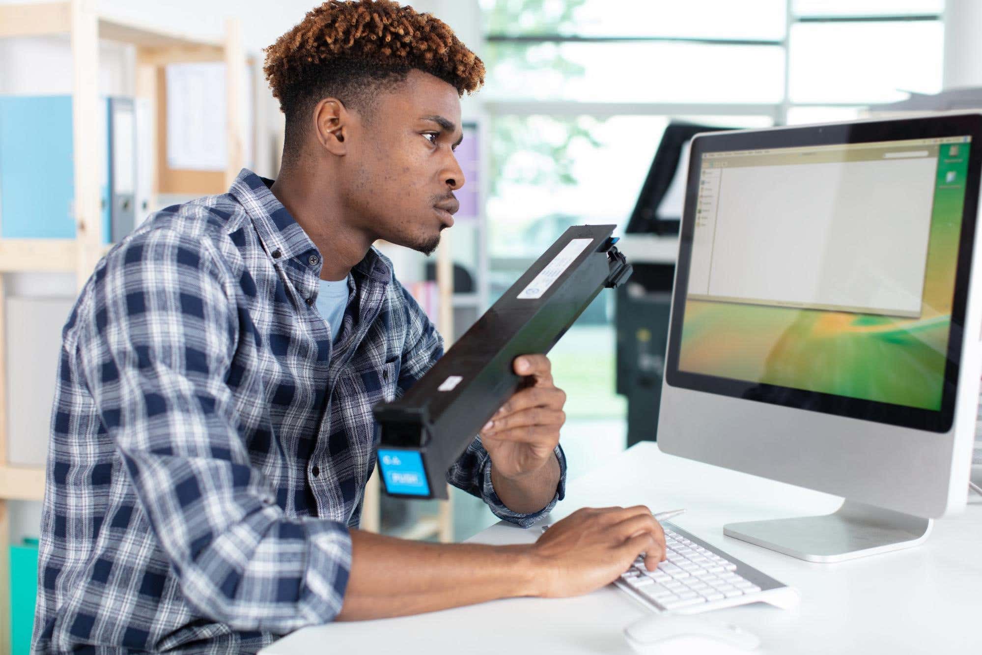 Man holding a print cartridge while sitting in front of a computer screen.